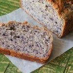 A loaf of Blueberry Almond Bread with a slice laying face up on parchemnet paper and a green background