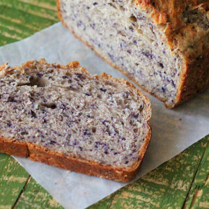 A loaf of Blueberry Almond Bread with a slice laying face up on parchemnet paper and a green background