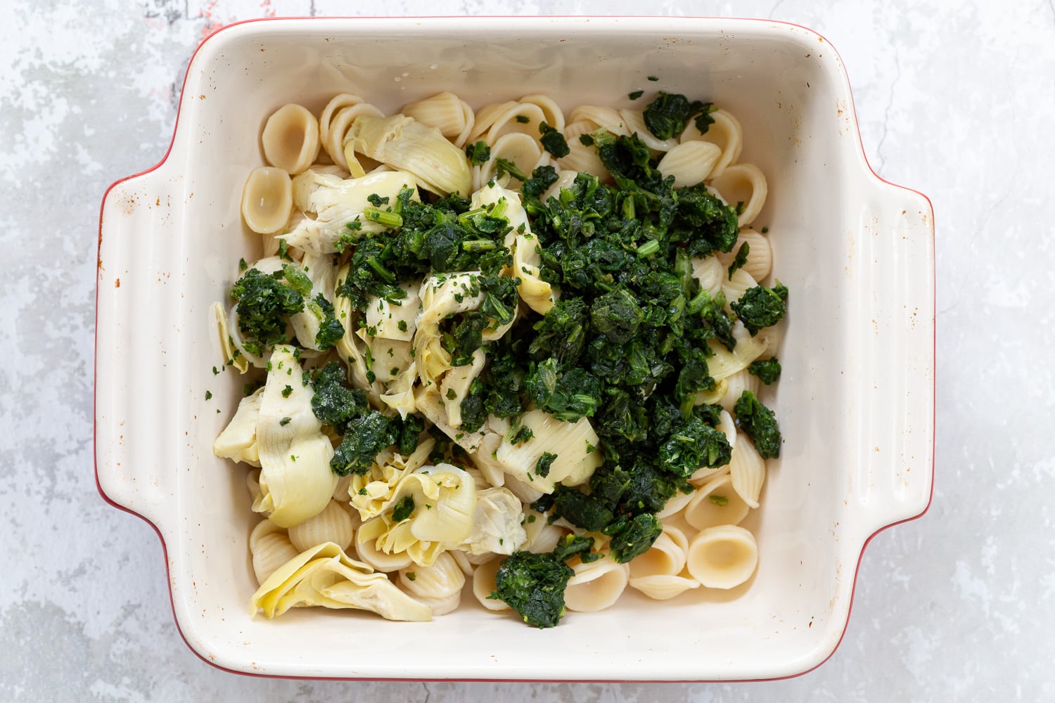 spinach pasta recipe overhead shot of a casserole dish with cooked shell pasta, spinach, and chopped artichokes