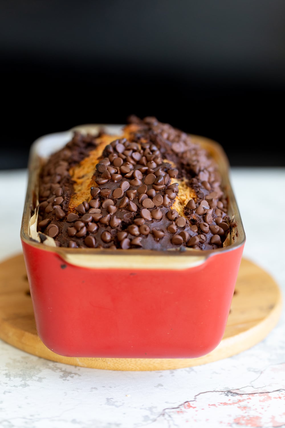 chocolate banana bread side view of a loaf of Vegan Chocolate Banana Bread topped with chocolate chips in a red ceramic loaf pan