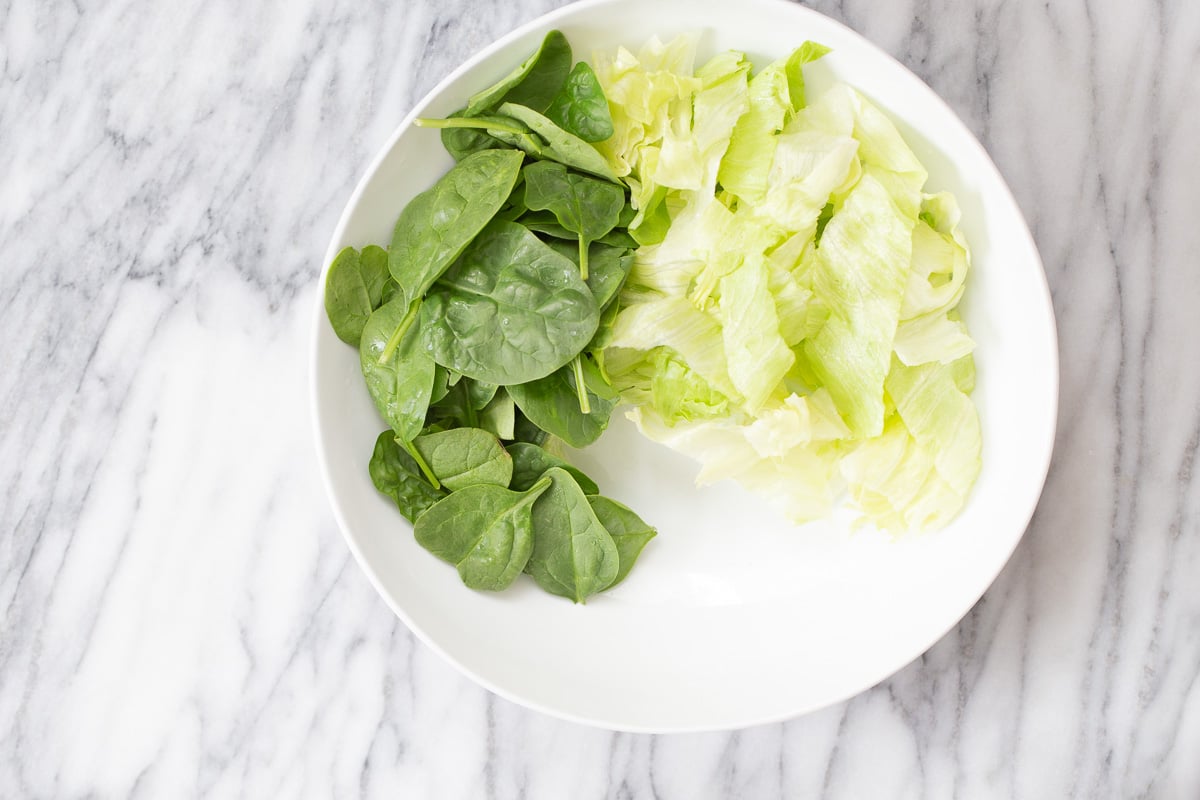 cauliflower salad spinach and iceberg salad being arranged in a white bowl to make cauliflower salad bowl