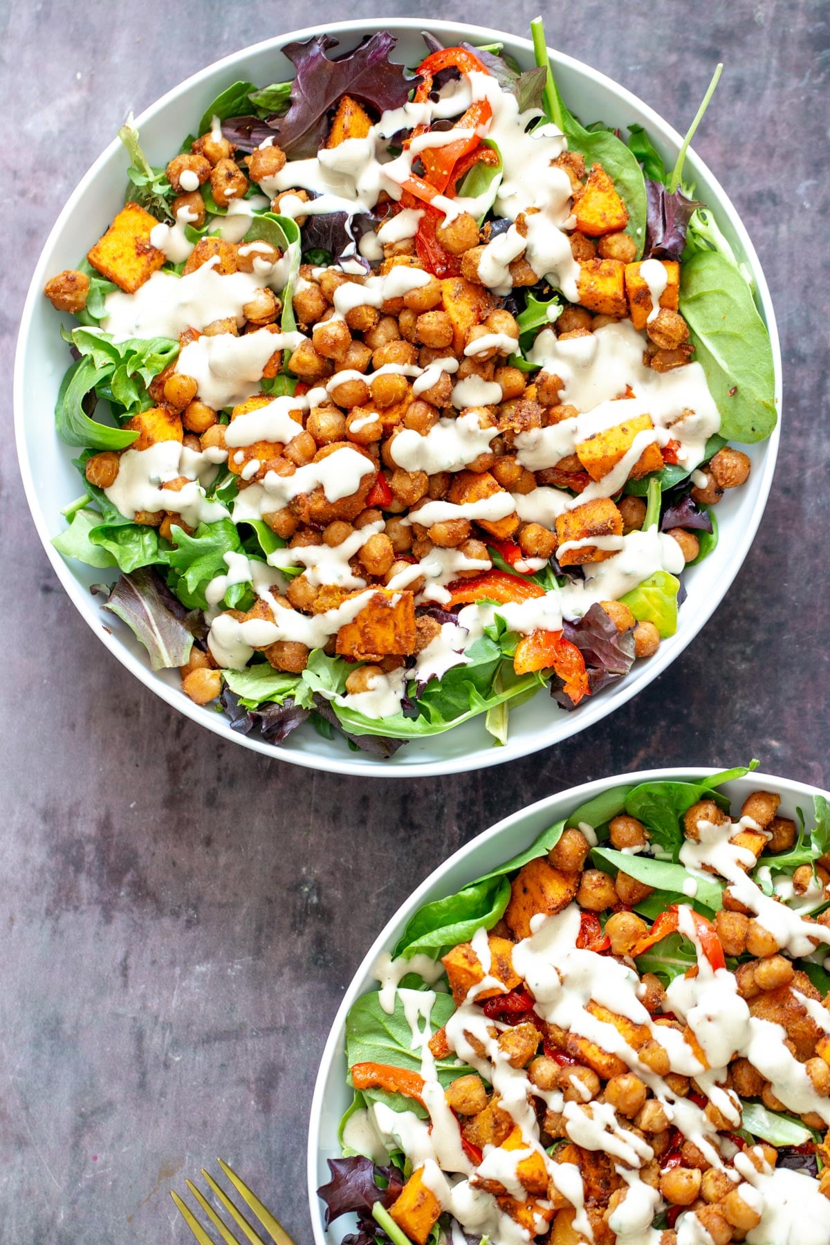 sweet potato chickpea salad two salad bowls with spring mix topped with roasted chickpeas, bell peppers and sweet potatoes and tahini dressing