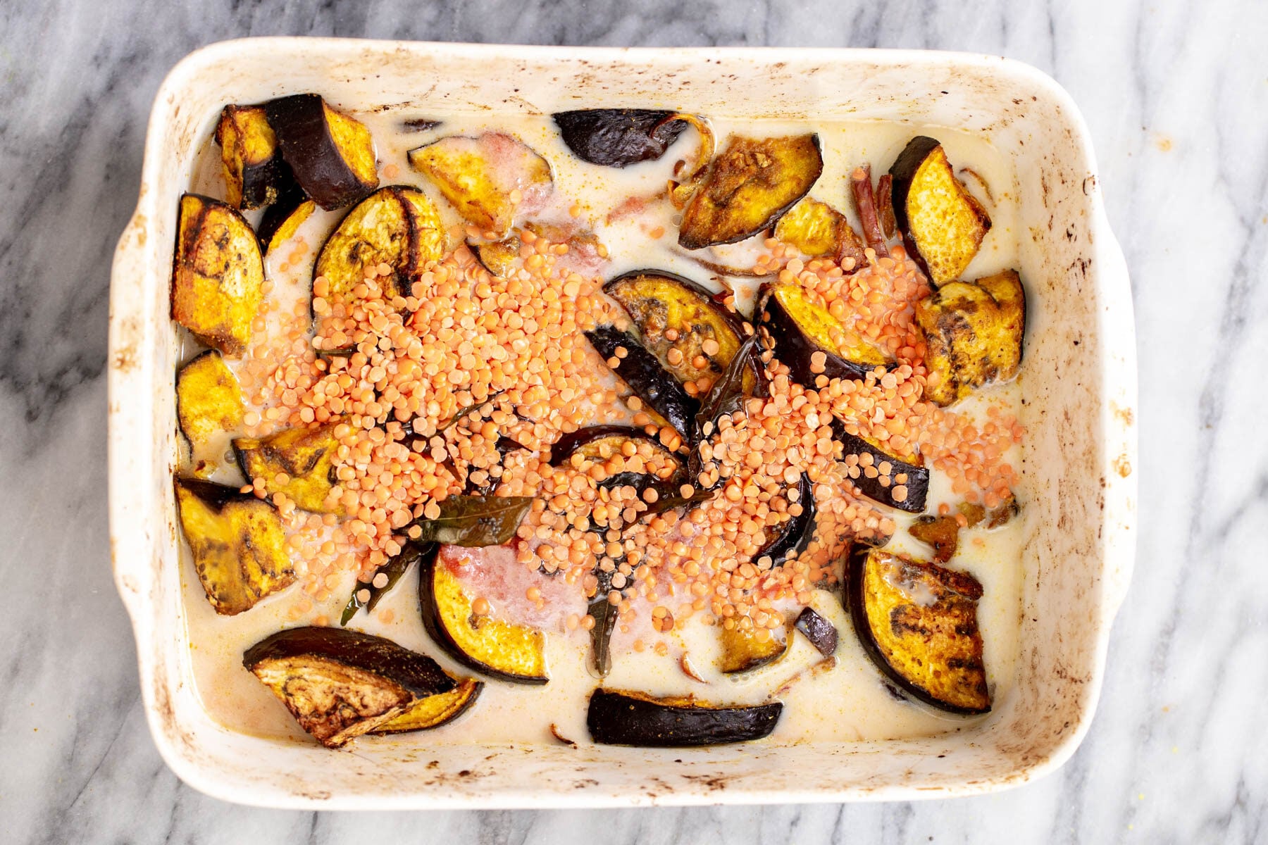 red lentils being sprinkled over eggplants in coconut milk in a white casserole dish