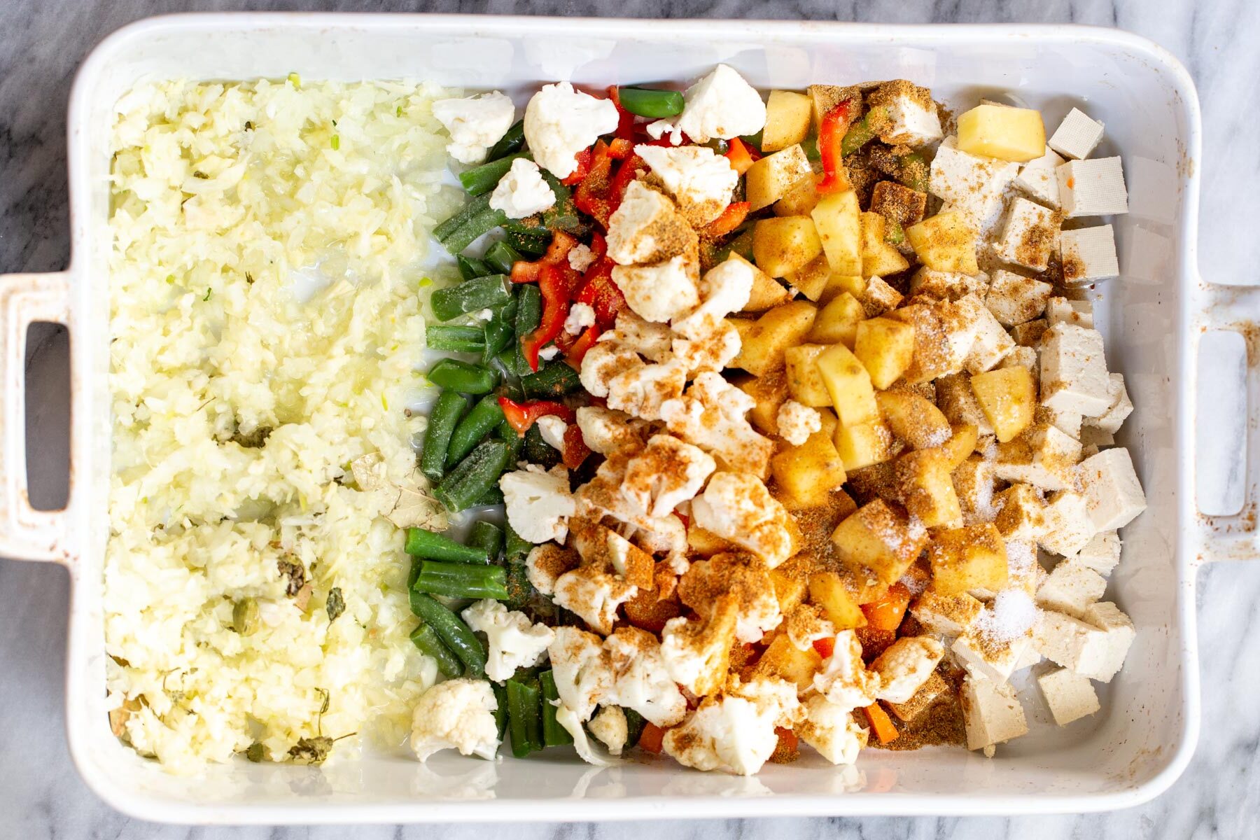 spices being sprinkled on top of veggies in a white casserole dish
