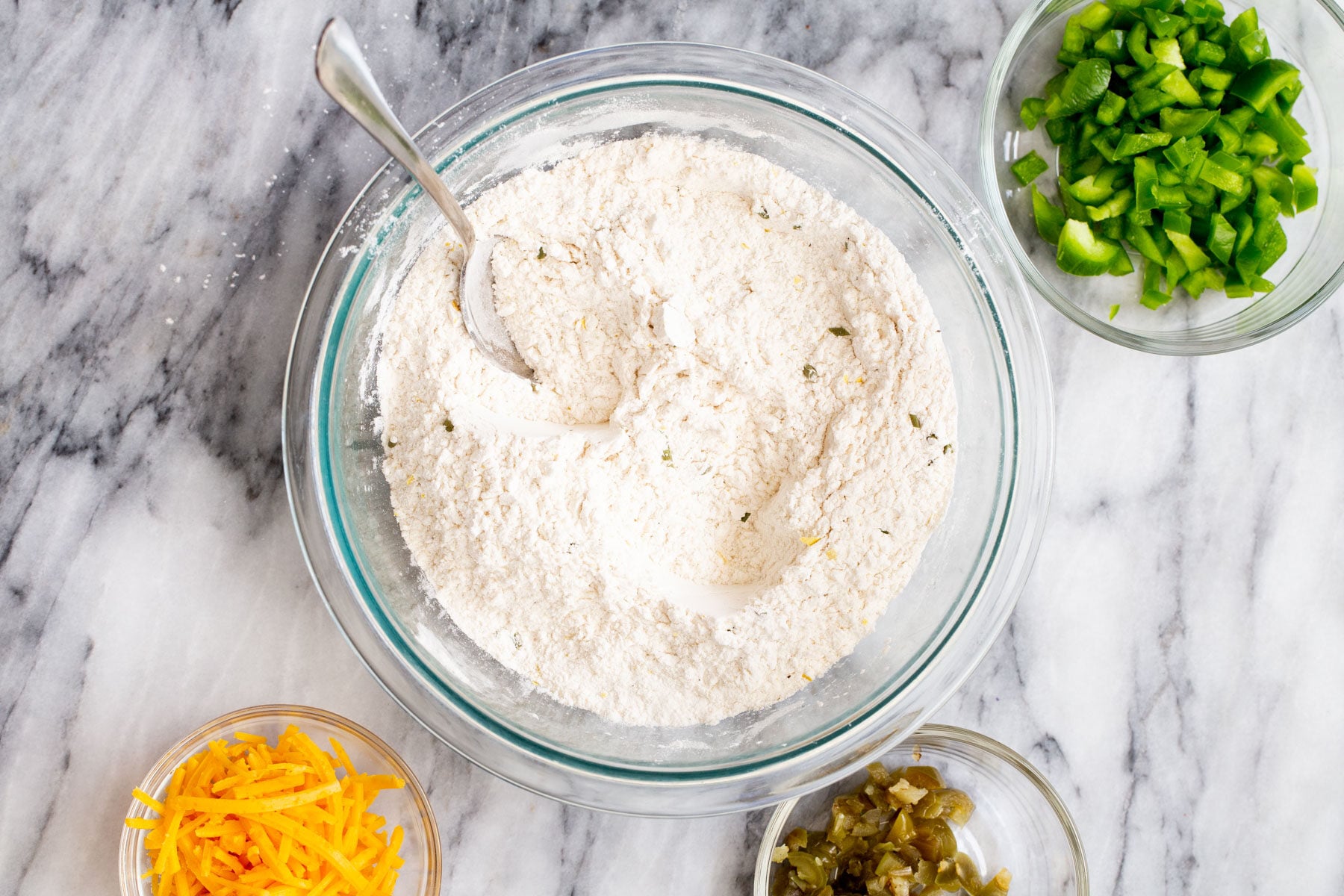 mixed dry ingredients in a glass bowl surrounded by ingredient dishes with jalapenos and cheddar