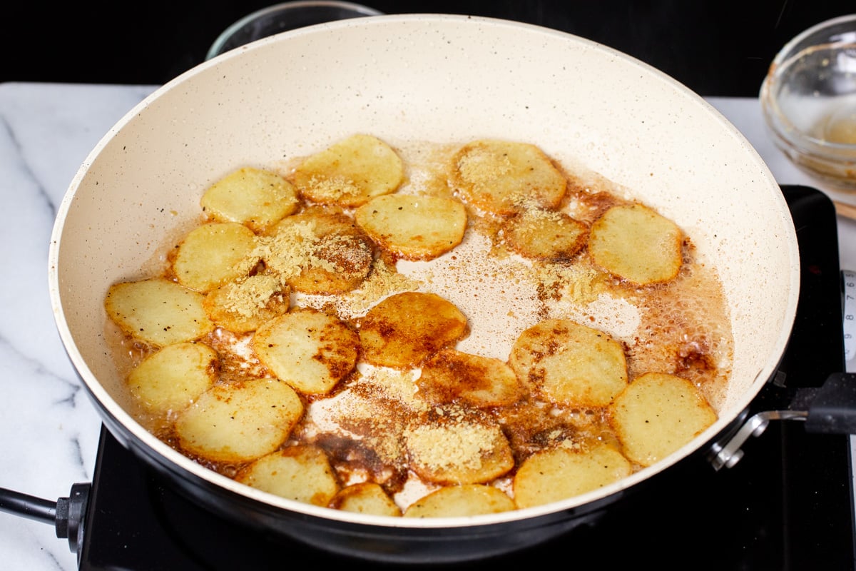 spices being added to fried sliced potatoes in a white pan 
