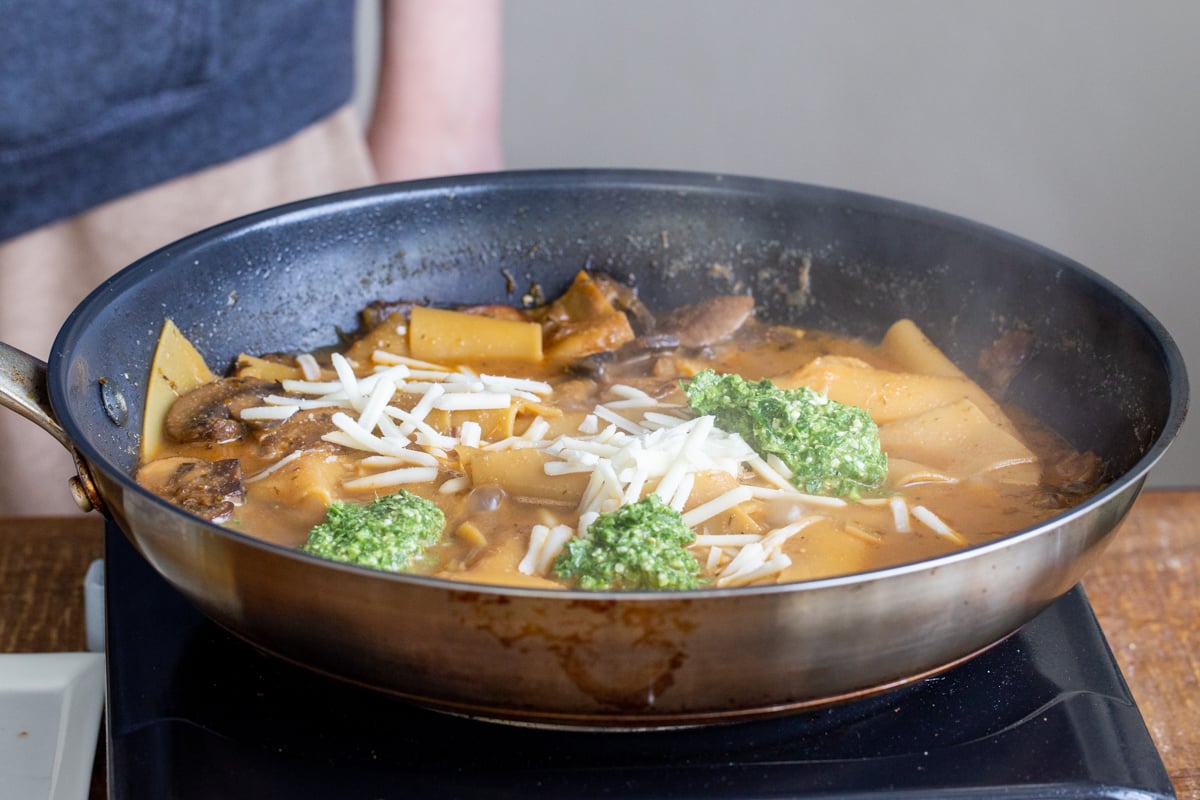 lasagna pasta simmering in a skillet topped with dollops of pesto and vegan cheese