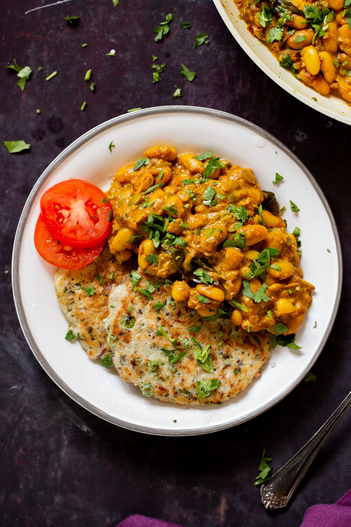 plate of spicy white bean stew with cilantro garnish, naan, and tomato