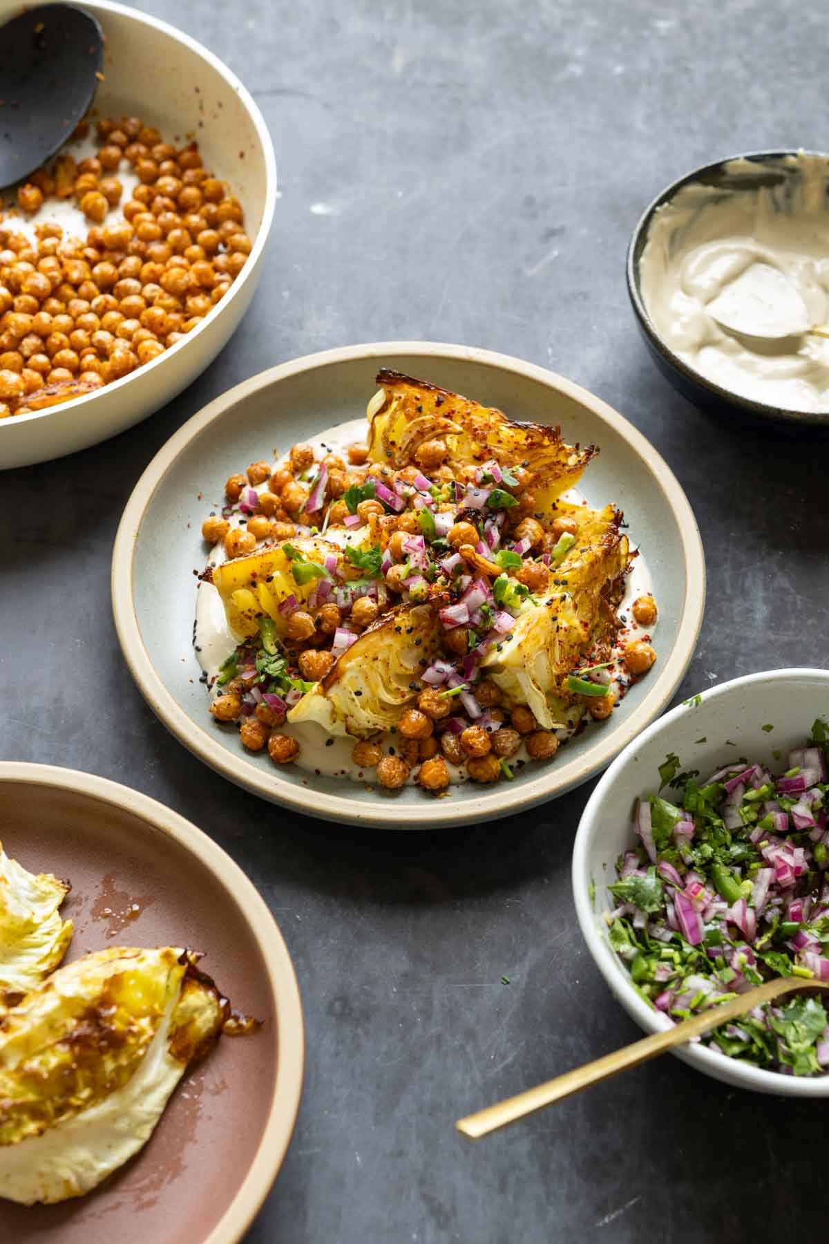 roasted cabbage wedges on a bed of tahini sauce with crispy chickpeas and gremolata surrounded by bowls of each component